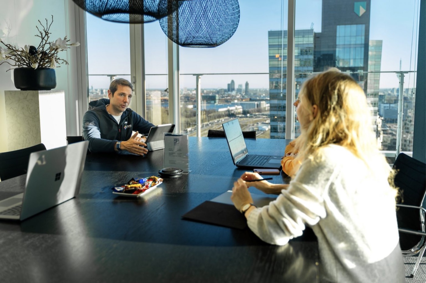 Two people talking across a meeting table with laptops, with a view over Amsterdam