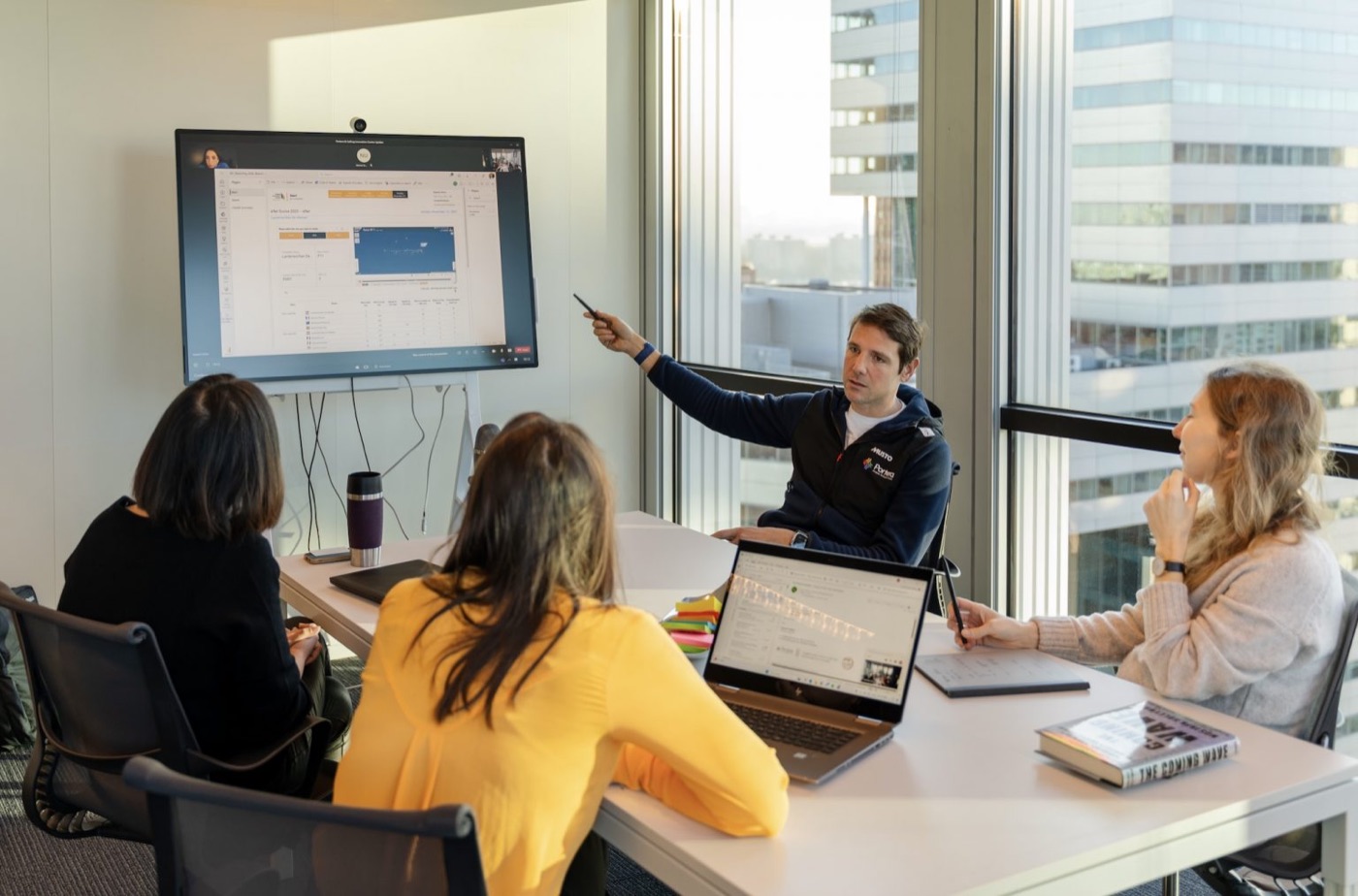 A presenter points at sailing performance data on a wall screen while four colleagues follow along